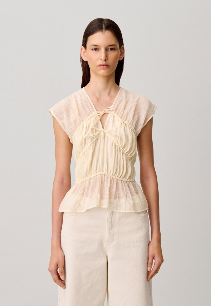 Giovane donna con lunghi capelli scuri che indossa una camicetta senza maniche beige strutturata con orlo a peplo e pantaloni crema a vita alta su sfondo uniforme.