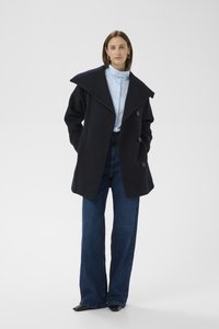 Tengerészkék gyapjú kabát nagy gallérral, kétsoros gombolású kialakítással, világoskék blúzzal és magas derekú, széles szárú kék farmernadrággal párosítva.