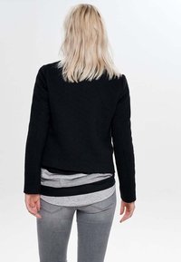Personne blonde portant une veste noire texturée, des chemises grises et noires superposées, et un jean gris, tourné le dos à un fond uni.