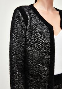 Svart och grå strukturerad cardigan med en fluffig finish, V-hals och två framfickor mot en vit bakgrund.