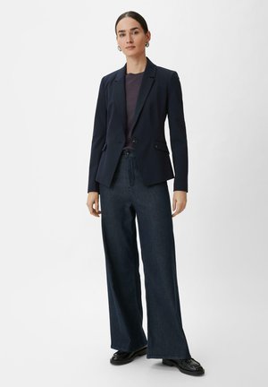 Navy blazer with a single button, lapels, and two side pockets, paired with loose-fitting dark denim trousers and black shoes.