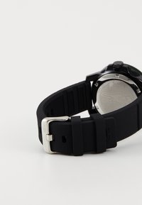 Bracelet de montre en silicone noir avec une boucle en métal argenté, présentant un dessous texturé et un logo détaillé sur le bracelet.