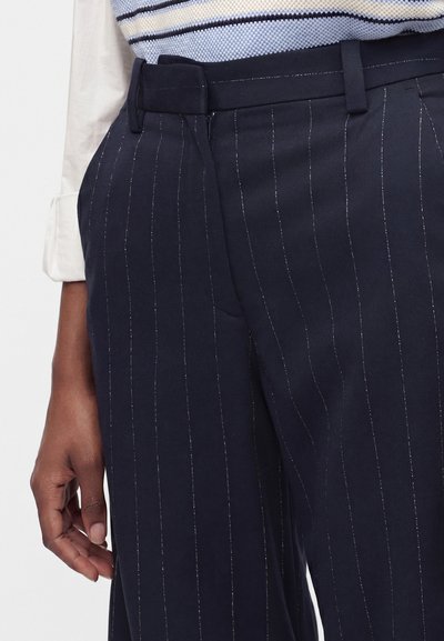 Gros plan d’une personne portant un pantalon bleu marine à fines rayures et une chemise blanche aux manches retroussées, main détendue sur le côté.
