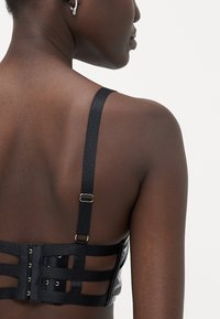 Črna strukturirana bralette s prilagodljivimi naramnicami, ki ima zanko in očesce za zapiranje ter izrezane detajle na hrbtu za dodatno estetiko.