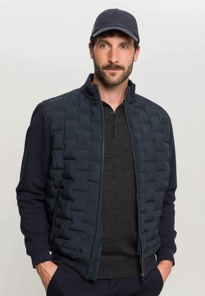 Vesta puffer bleu marin cu textură matlasată, închidere cu fermoar și guler înalt, asociată cu un tricou polo închis la culoare. Completată cu o șapcă asortată.