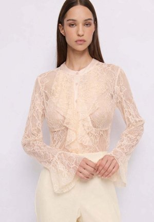 Donna con lunghi capelli castani che indossa una camicetta di pizzo beige trasparente con volant e maniche svasate, abbinata a pantaloni chiari.