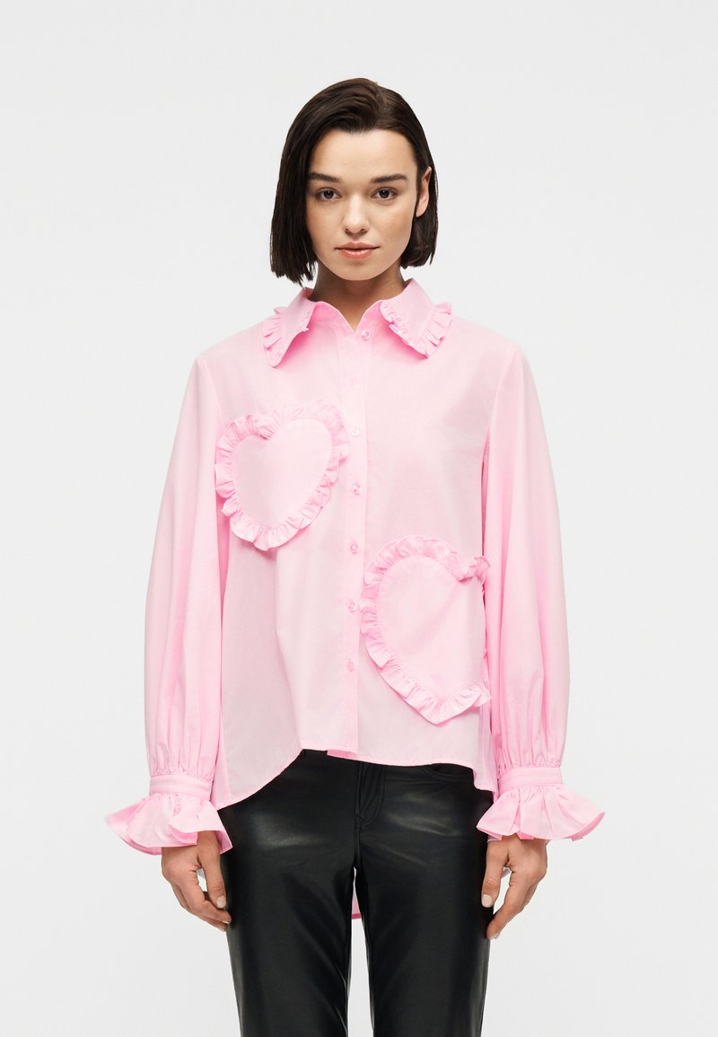 Γυναίκα που φοράει μια φαρδιά ροζ πουκάμισο με κουμπιά και βολάν σε σχήμα καρδιάς στο μπροστινό μέρος και βολάν στις μανσέτες, συνδυασμένο με μαύρο παντελόνι, σε ουδέτερη στάση.