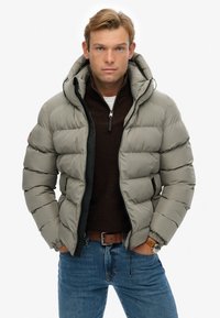 En blond mand iført en lys grå pufferjakke, brun kvart-zip sweater, blå jeans, brun læderbælte og armbåndsur, stående med hænderne i lommerne.