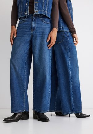 Dwie osoby noszące szerokie niebieskie dżinsy i czarne ostro zakończone buty, stoją blisko siebie na jednolitym tle.