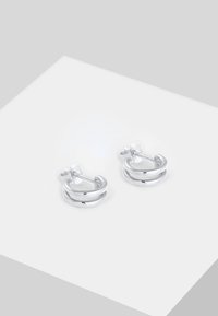 Paire de petites boucles d'oreilles créoles argentées à double bande avec fermoir à tige sur une surface blanche inclinée.