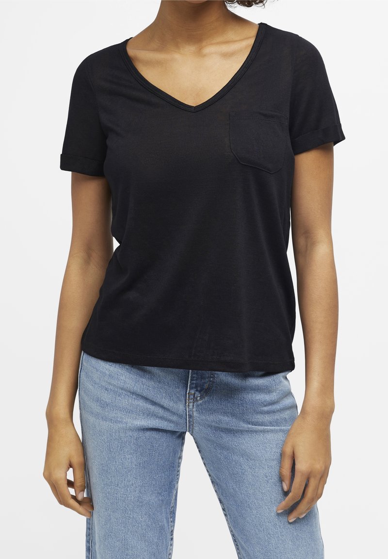 Femme portant un T-shirt noir à manches courtes avec col en V et poche poitrine, et un jean bleu clair, montrée des épaules aux hanches.