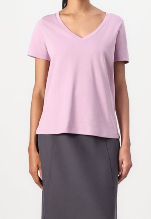Frau, die ein hellviolettes T-Shirt mit V-Ausschnitt und kurzen Ärmeln sowie einen dunkelgrauen Rock trägt, steht vor einem einfarbigen Hintergrund.