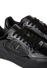 Crne tenisice s gornjom od lakirane kože, teksturalnim detaljima, vezanjem i akcentom logotipa sa strane.