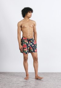 Homme torse nu avec des cheveux bouclés, portant un short de bain floral bleu marine, debout pieds nus sur un sol en pierre grise, regardant sur le côté.
