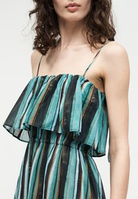 Γυναίκα που φοράει αμάνικο, αέρινο φόρεμα με κάθετες ρίγες σε τιρκουάζ, μαύρο, άσπρο και καφέ, με λεπτά τιραντάκια στους ώμους.