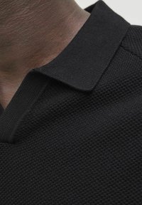 Polo nero realizzato in un tessuto testurizzato con un motivo intrecciato. Il colletto è piatto e leggermente strutturato, in armonia con il design generale.