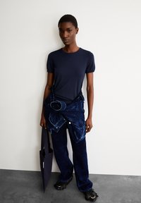 Marineblauw aansluitend t-shirt met donkere spijkerbroek, opgesierd met een oversized spijkerjack dat om de taille is gestrikt. Zwarte fringeschoenen en een zwarte totebag.