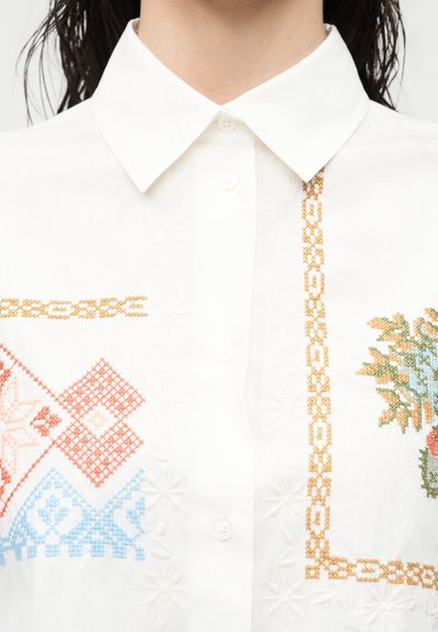Λευκό πουκάμισο με γιακά, διακοσμημένο με πολύχρωμα σχέδια κεντήματος σταυροβελονιάς, που περιλαμβάνουν λουλούδια, γεωμετρικά σχήματα και κατακόρυφο διακοσμητικό περιθώριο κοντά στα κουμπιά.