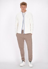 Giacca bianca con zip e dettagli blu, abbinata a pantaloni jogger marroni. L'outfit presenta texture morbide e una vestibilità rilassata. Le sneakers bianche completano il look.