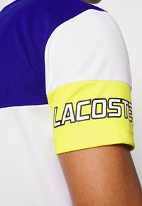 Personne portant une chemise color block avec une manche bleue, blanche et jaune, affichant un texte noir en gras « "LACOSTE" » sur la bande jaune.