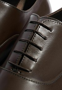 Scarpe in pelle marrone con una finitura liscia, caratterizzate da un design con lacci e dettagli finemente cuciti lungo i lati.