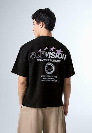 Muškarac u crnoj majici s natpisom "SUBVISION Below the Surface" i grafikom zvijezda, upareno s bež hlačama, okrenut leđima kameri.