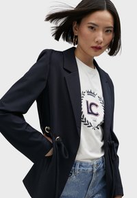 Navy blazer with a tailored design, gold hardware, and drawstring accents. Worn over a white T-shirt featuring a graphic logo and blue denim jeans.