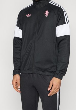 Homme portant une veste et un pantalon de survêtement Adidas noirs avec des bandes blanches et des logos roses Adidas et cheval cabré.