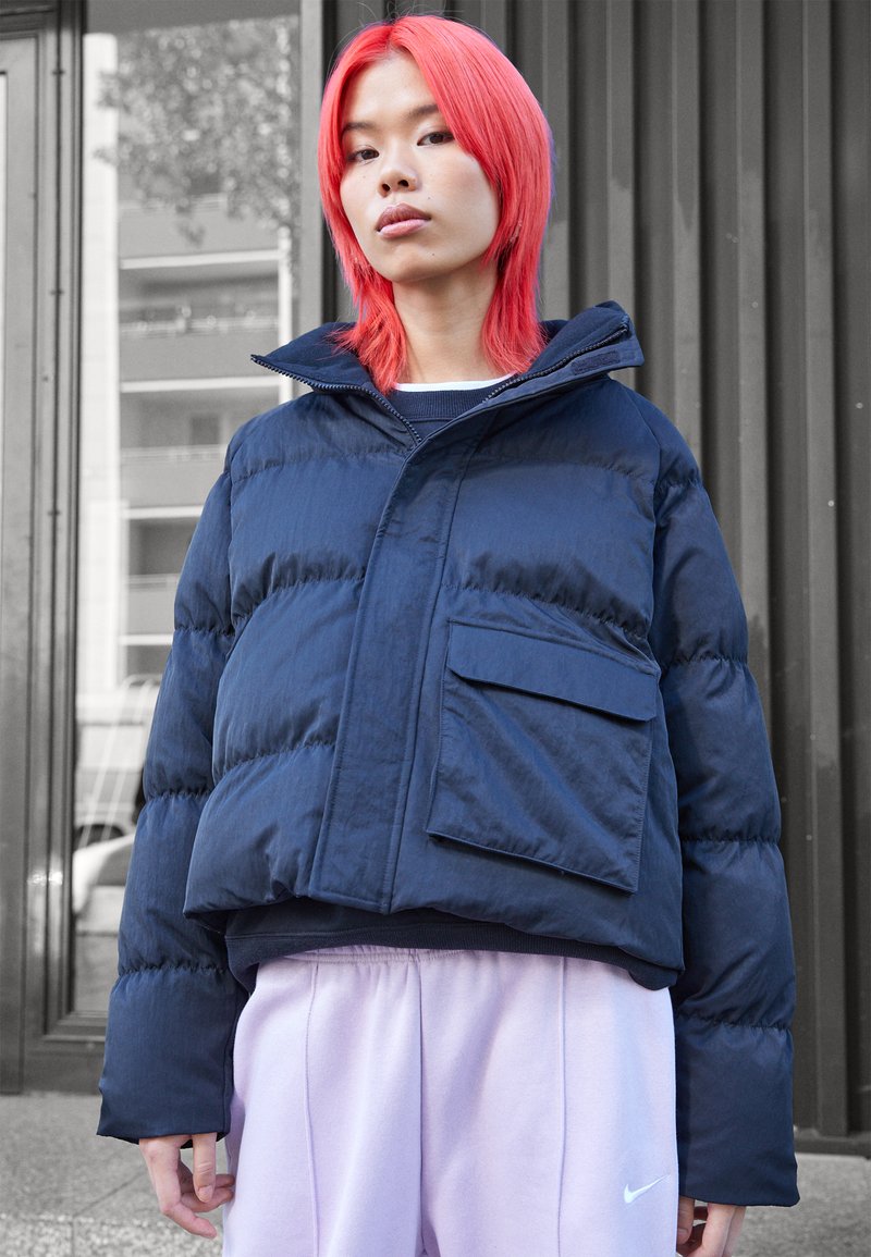 Doudoune navy avec un col haut, une grande poche à l'avant et une texture matelassée, associée à un pantalon de survêtement rose clair. Finition en tissu lisse.