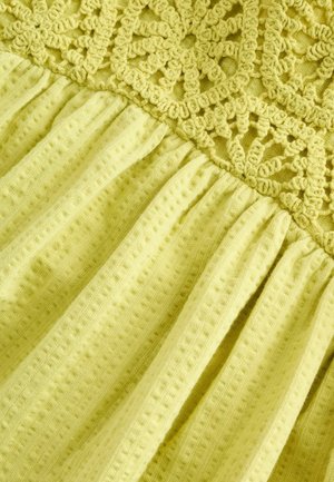 Tessuto lavorato a maglia giallo con superficie strutturata e dettagli decorativi in crochet nella parte superiore, accompagnato da pieghe arricciate nella parte inferiore.