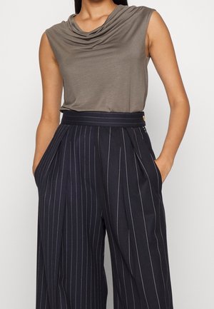 Donna che indossa una maglia senza maniche grigia drappeggiata infilata in pantaloni a vita alta neri a righe sottili a gamba larga, con le mani nelle tasche.