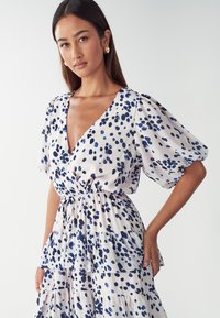 Robe florale en tissu léger avec un décolleté en V profond, des manches bouffantes et des volants superposés. Présente un motif de pois bleus et marins sur un fond clair.