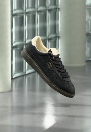 Sneaker nero low-top con fodera interna beige e suola marrone, che fluttua sopra un pavimento in cemento lucidato in uno spazio industriale moderno.