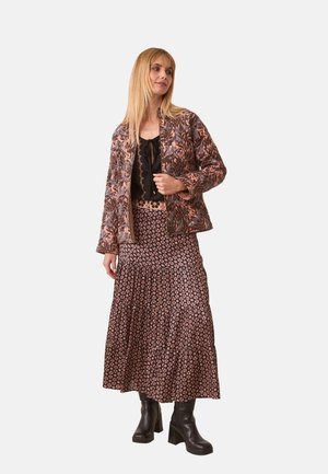 Mujer vestida con chaqueta acolchada estampada, top negro, falda larga con flores y botas negras de tacón, de pie sobre un fondo liso de color gris claro.