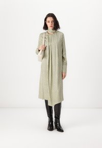 Grønt mønstrete kjole med lange ermer og høy krage, kombinert med høye svarte lærstøvler og en liten beige veske.