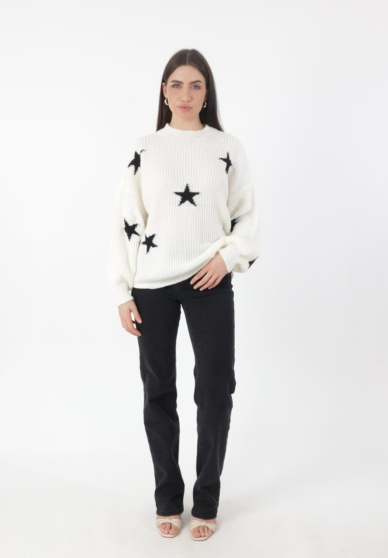 Overgroot wit gebreid sweater met zwarte sterpatronen, gecombineerd met zwarte broek en lichtgekleurde sandalen. Eenvoudig ontwerp met een ontspannen pasvorm.