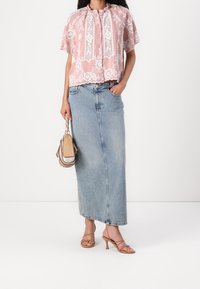 Vaaleanpunainen kirjailtu pusero, jossa on lyhyet hihat ja napitettava edusta, yhdistetty farkkumaksihameeseen. Pitelee pientä vaaleanruskeaa käsilaukkua.