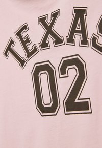 Lichtroze T-shirt met vetgedrukte zwarte tekst die "TEXAS" boven het nummer "02" in een boog weergeeft, met een zachte textuur en klassiek ontwerp.