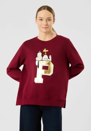 Woman wearing a burgundy sweatshirt with Snoopy and Woodstock reclining on a large gold letter P, standing with hands behind her back.