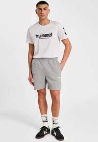 T-shirt blanc à col rond avec le logo "hummel" noir, associé à un short de sport gris. Le modèle porte des baskets noires et des chaussettes blanches rayées.