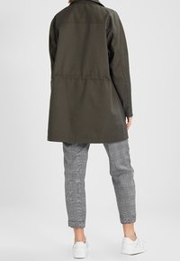 Olivgrön lång kappa med rak skärning, har en söm i ryggen, kombineras med grå rutiga byxor och vita sneakers.