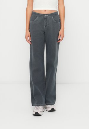 Jeans grigi ampi con vita alta, caratterizzati da dettagli cuciti, tasche anteriori e una texture liscia. Abbinati a sneaker di colore chiaro.