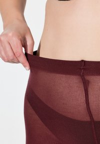 Burgundowe przezroczyste rajstopy z teksturowanym pasem, pokazujące zbliżenie na dłoń chwytającą górną część. Tkanina wydaje się lekka i rozciągliwa.