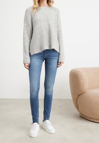 Suéter de punto gris con un corte holgado, dobladillo asimétrico, combinado con jeans ajustados azules y zapatillas blancas. Textura suave y diseño casual.