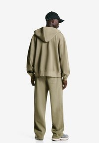 Olive green hoodie with a drawstring hood, relaxed fit, and ribbed cuffs, paired with matching loose-fit pants and gray sneakers.