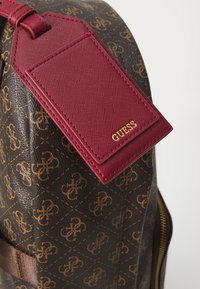 Sac à dos marron à motifs avec une étiquette en cuir bordeaux portant le logo doré "GUESS", en matériau texturé et avec un accent doré sur la fermeture éclair.