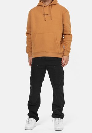 Tan cotton hoodie with front pocket, featuring a black logo. Paired with black cargo pants with pockets and distressed detailing, and white sneakers.