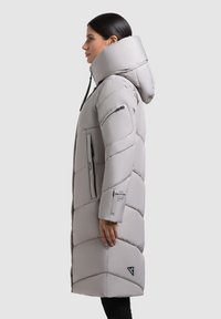 Abrigo tipo puffer en gris claro con patrón acolchado diagonal, cuello alto y bolsillos con cremallera. Incluye capucha y detalle de logo en el costado.