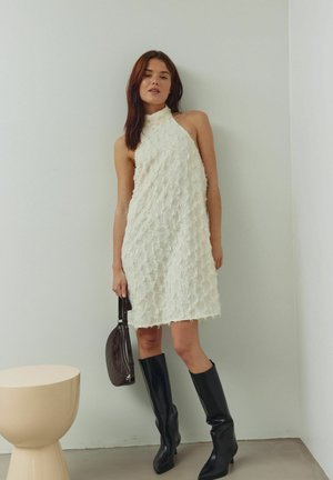 Balta haltera stila kleita ar teksturētu bārkstīm, saskaņota ar melniem ceļgalu augstiem zābakiem. Modele tur tumši brūnu rokassomiņu.