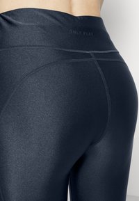 Tummansiniset leggingsit, joissa on sileä, kiiltävä pinta, korkeavyötäröinen ja litteillä saumoilla mukavuutta varten. Brändin logo on hienovaraisesti sijoitettu yläosaan.